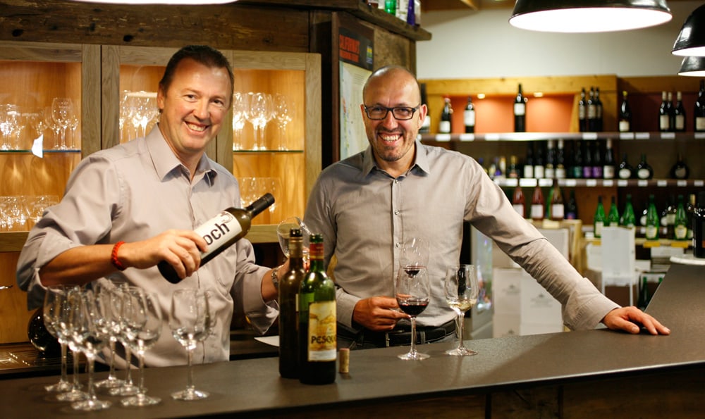 Zwei Männer lächeln, einer hält eine Weinflasche mit der Aufschrift „bach“, der andere ein Glas Rotwein.