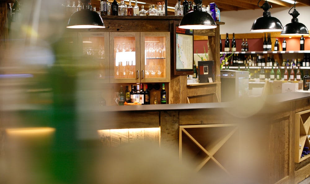 Holzbar mit Weingläsern, Flaschen und drei schwarzen Hängelampen in einem Weingeschäft.